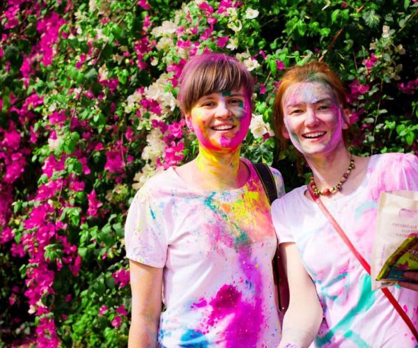 Two travelers smiling, covered in vibrant Holi colors, standing in front of a bougainvillea background during a group tour in India.