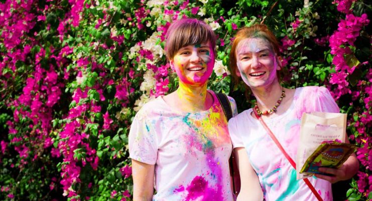 Two travelers smiling, covered in vibrant Holi colors, standing in front of a bougainvillea background during a group tour in India.