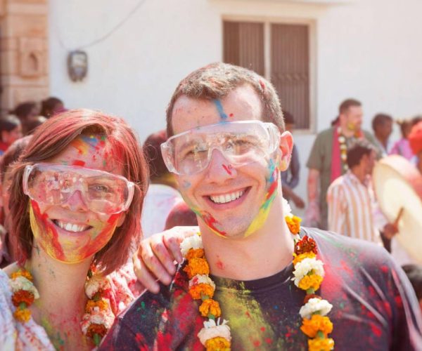 Tourist couple enjoying Holi celebration in India, covered in colorful powder and wearing marigold garlands.