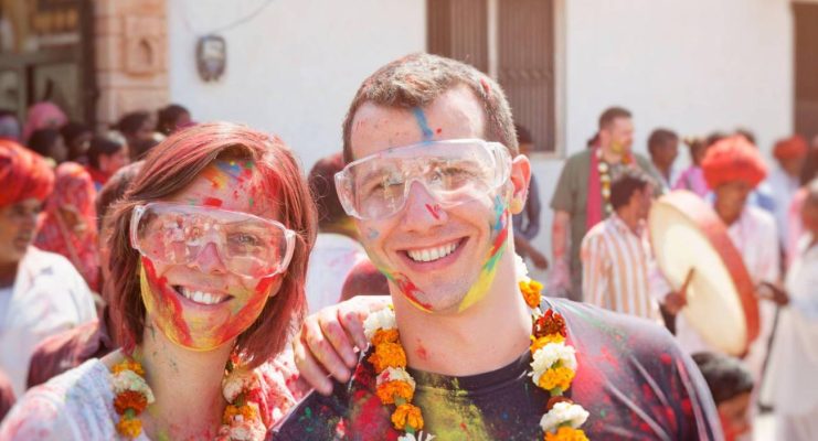 Tourist couple enjoying Holi celebration in India, covered in colorful powder and wearing marigold garlands.